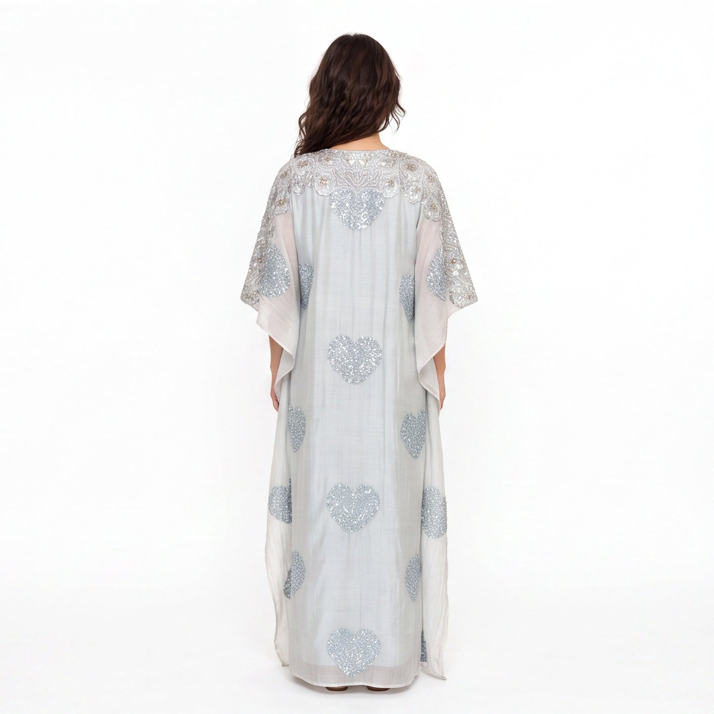A woman viewed from the back wearing a long sheer kaftan dress in pale blush pink with silver heart-shaped sequin embroidery, set against a plain white studio background.