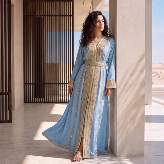 A woman leaning against a stone pillar in a flowing sky blue handmade Moroccan kaftan with ornate gold embroidered front panel, belt and cuffs.