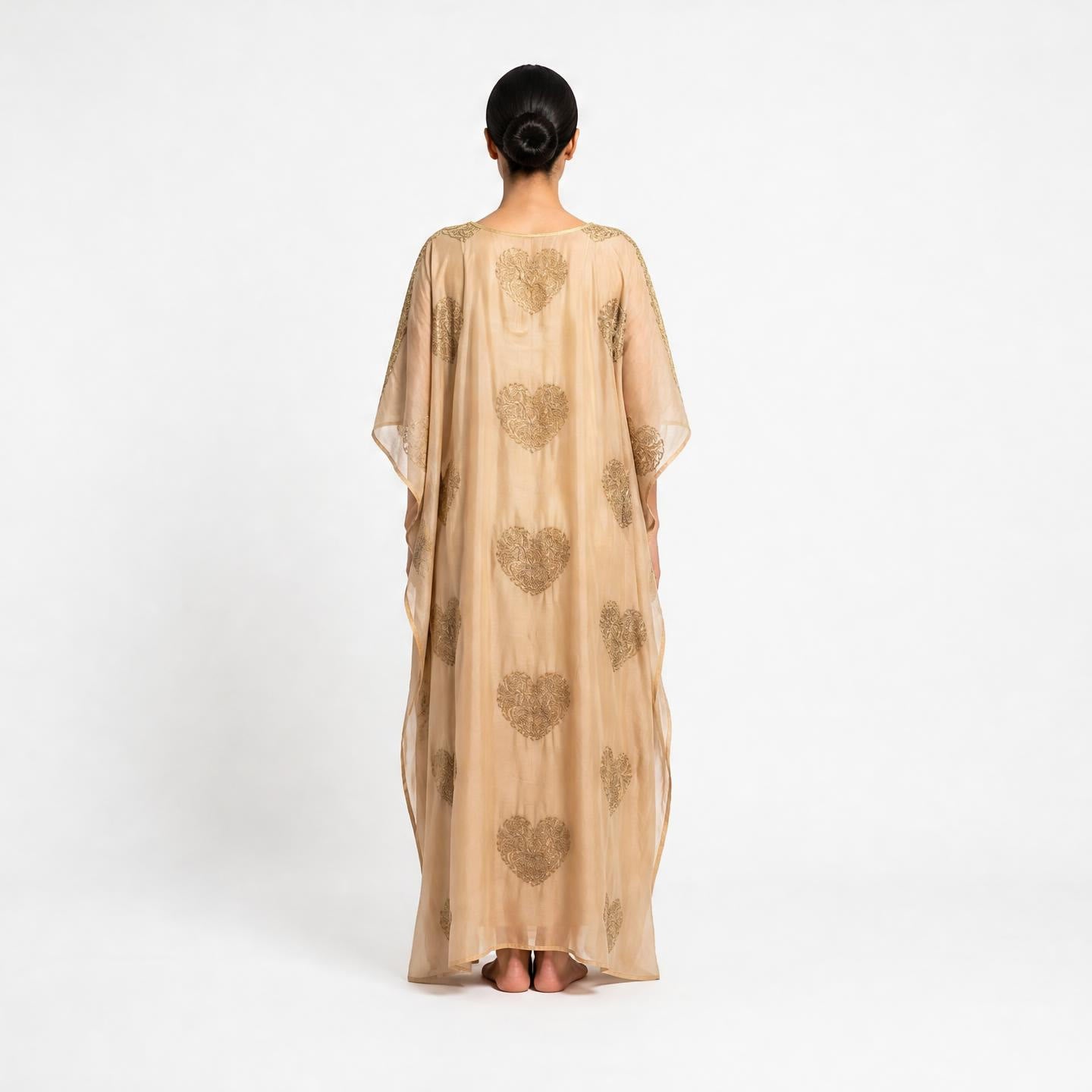 A woman viewed from behind wearing a flowing beige kaftan dress with metallic gold heart patterns, set against a plain white studio background.