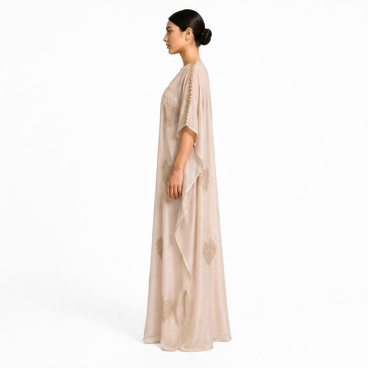 A woman in profile wearing a floor-length beige caftan with gold floral embroidery and sheer draped sleeves, photographed against a plain white studio background.