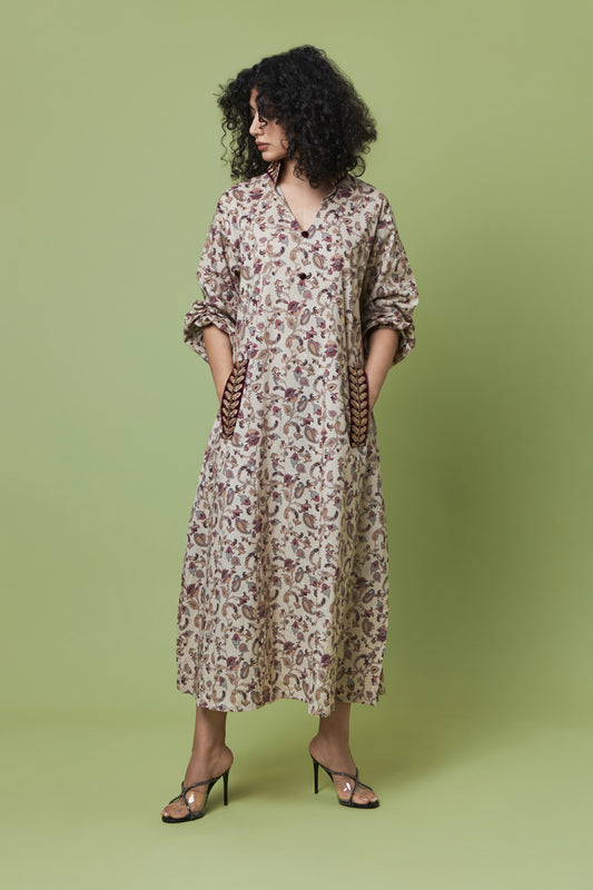 A woman modeling a long-sleeved, knee-length floral dress with a V-neck and side pockets, standing against a solid sage green studio background.