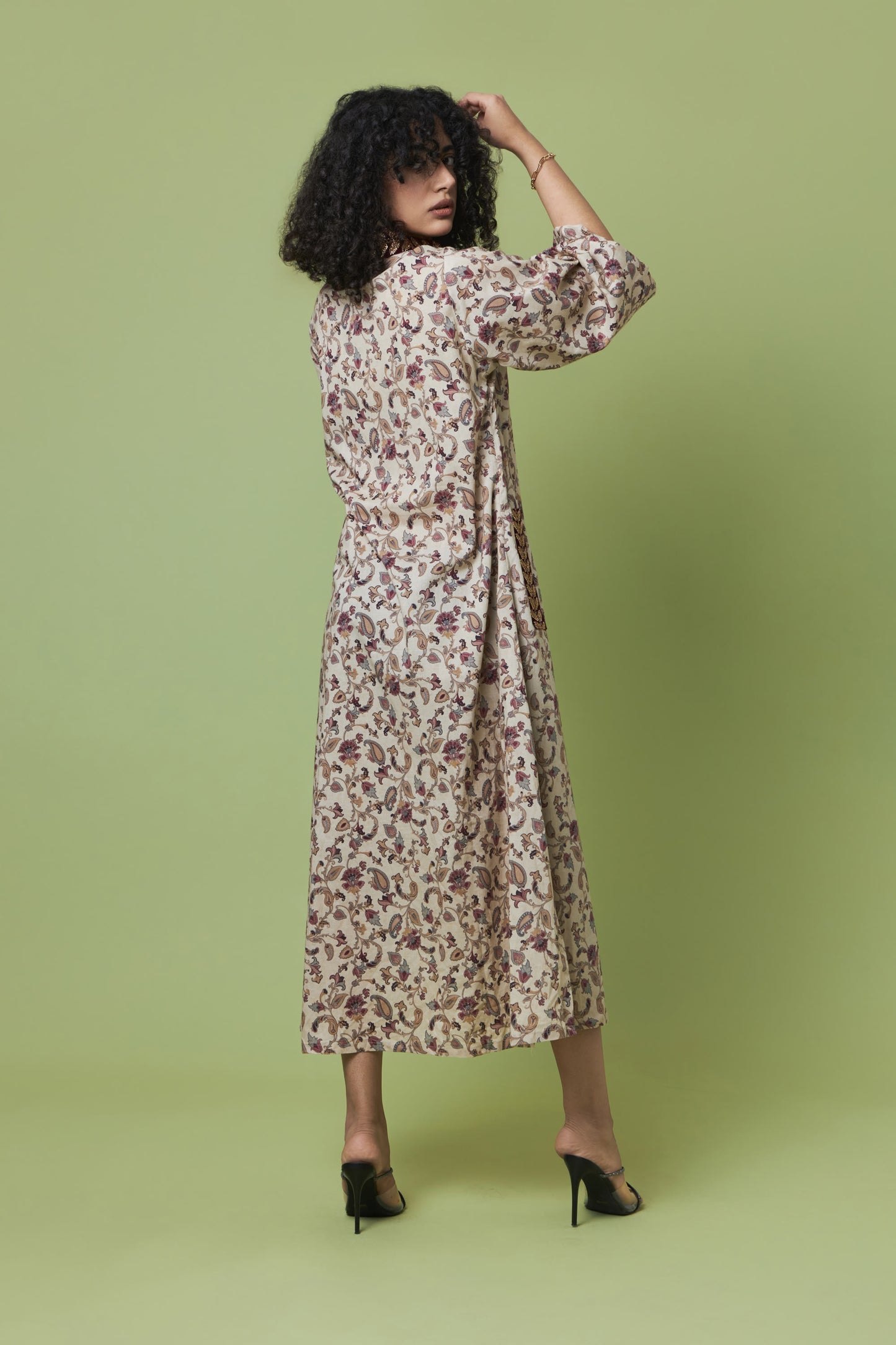A woman with curly black hair is shown from the back wearing a long-sleeved midi dress with a dense floral print in brown and muted red on a cream background. She is posing against a solid light green studio backdrop.