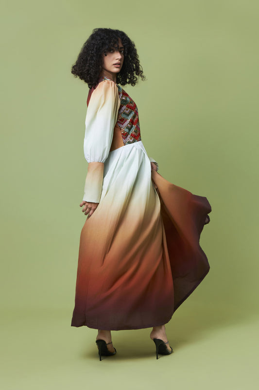 A model with curly black hair is shown from the back wearing a long-sleeved maxi dress with a white-to-brown ombre skirt and a patterned bodice, set against a solid olive green studio background.