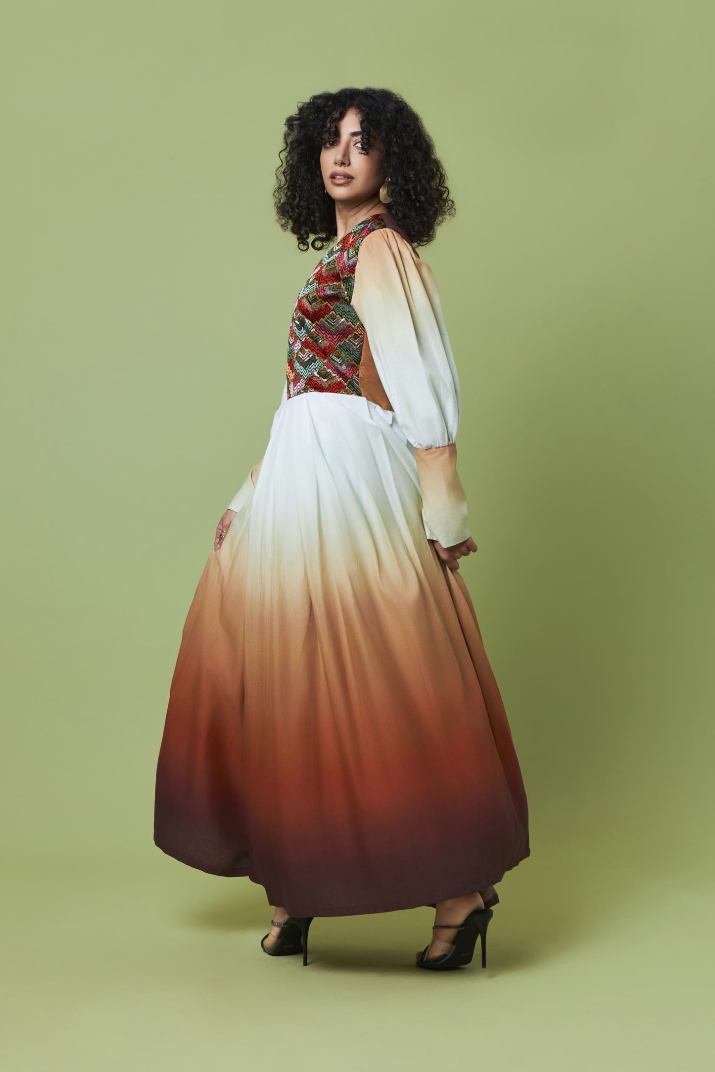A woman modeling a long maxi dress with a gradient design, featuring a multicolor patterned bodice, white upper skirt, and rust-orange to dark brown ombre lower skirt, set against a solid sage green background.