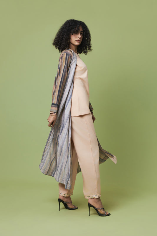 A model with curly black hair is shown from a side-back angle wearing a beige two-piece suit with a striped open-front cardigan and black high heels, set against a solid sage green studio background.