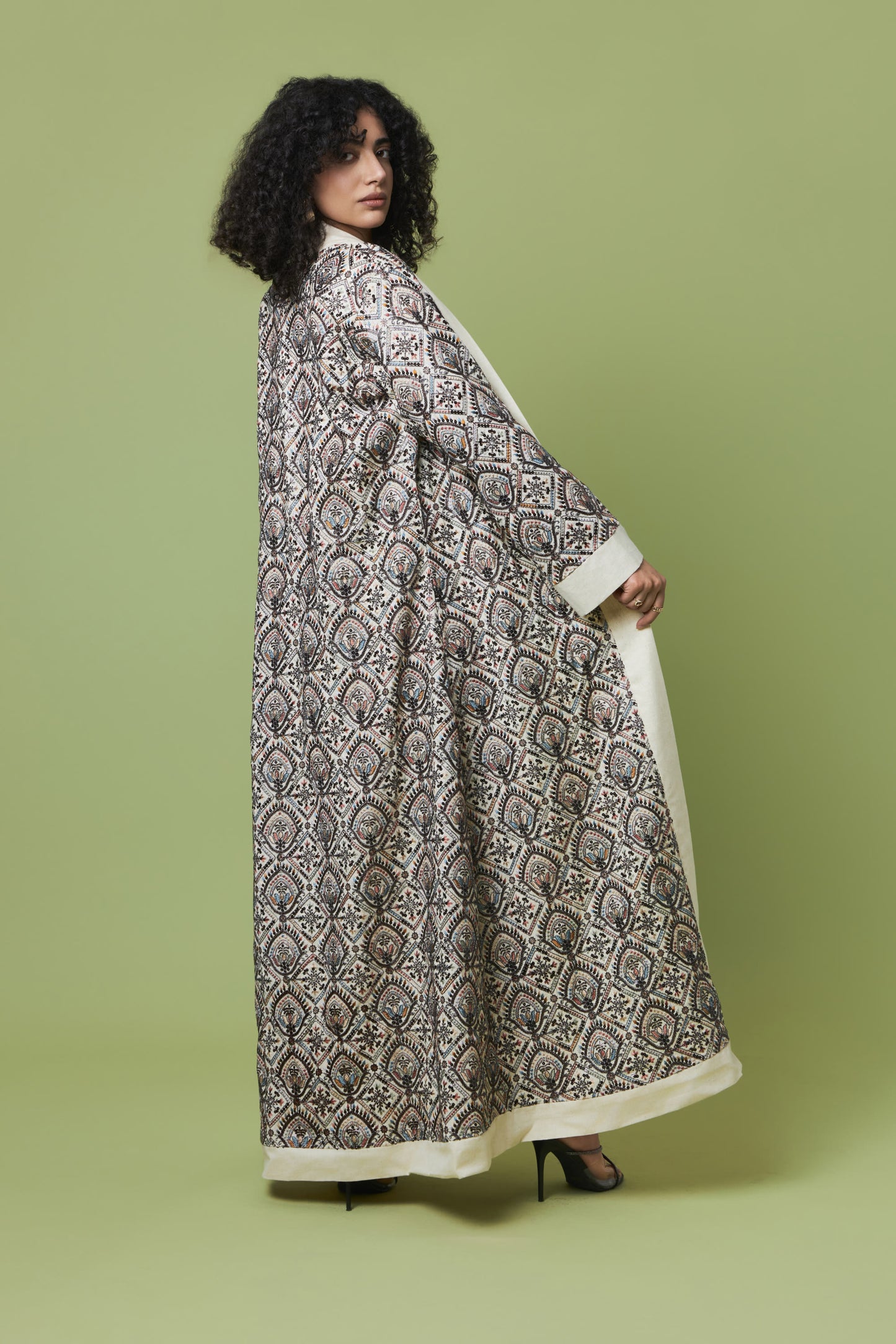 A woman modeling a long, flowing kaftan-style dress with an intricate black, gray, and beige floral pattern and solid cream borders, set against a solid sage green studio background.
