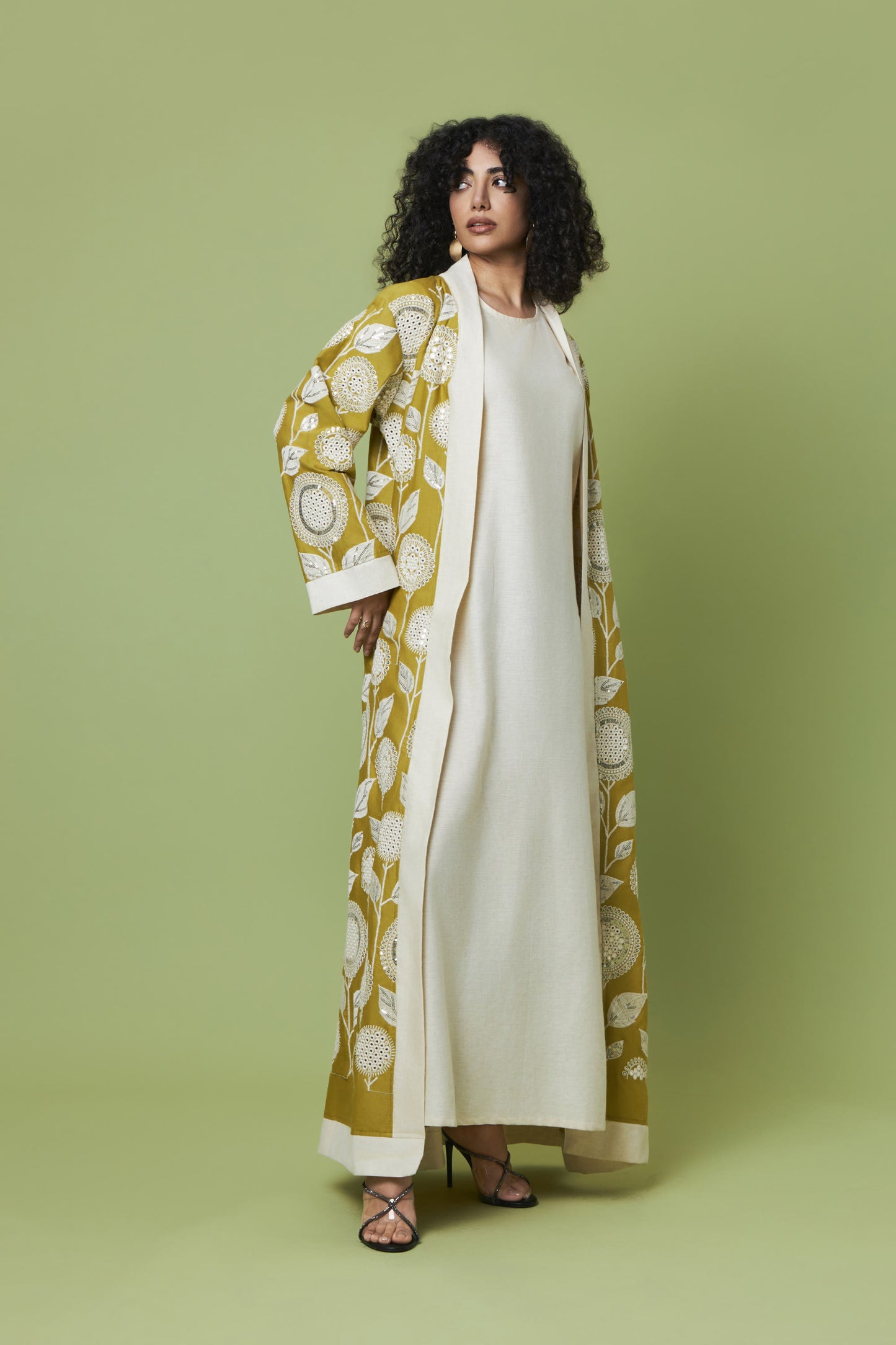 A woman modeling a long cream-colored dress with a matching open-front abaya featuring a mustard yellow and cream botanical pattern, set against a solid sage green studio background.