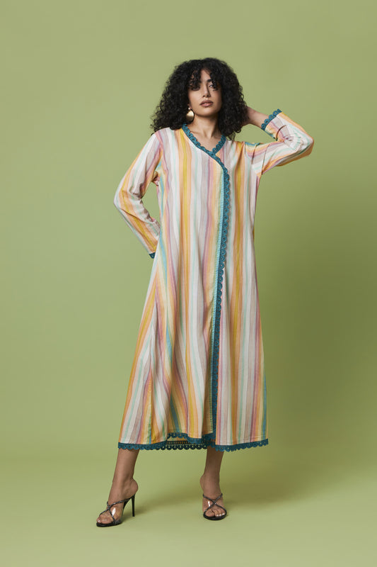 A woman modeling a long-sleeved, midi-length kaftan dress with vertical stripes in yellow, orange, pink, and white, accented with teal trim, set against a solid light green studio background.