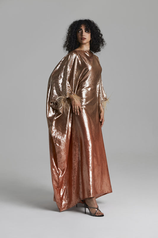 A model wearing a floor-length, metallic rose gold gown with wide draped sleeves and feathered cuffs, standing against a neutral gray studio background.