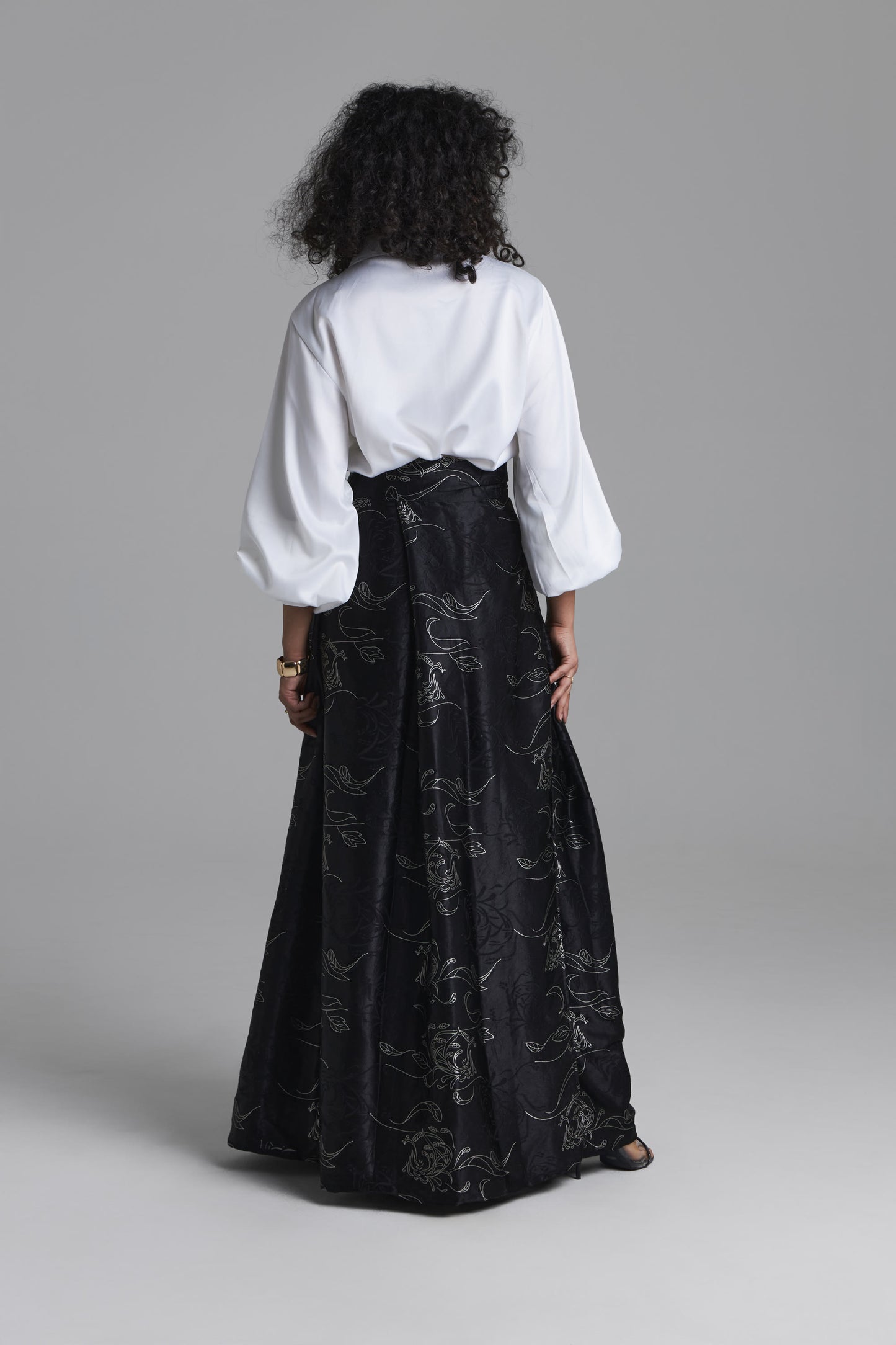 A model shown from the back wearing a white blouse with voluminous three-quarter sleeves and a long black skirt featuring a subtle tonal floral pattern, set against a solid gray studio background.