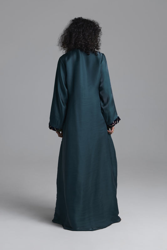 A person viewed from the back wearing a long dark teal satin-like robe or dress with long sleeves and ruffled cuffs, standing against a plain light gray studio background.