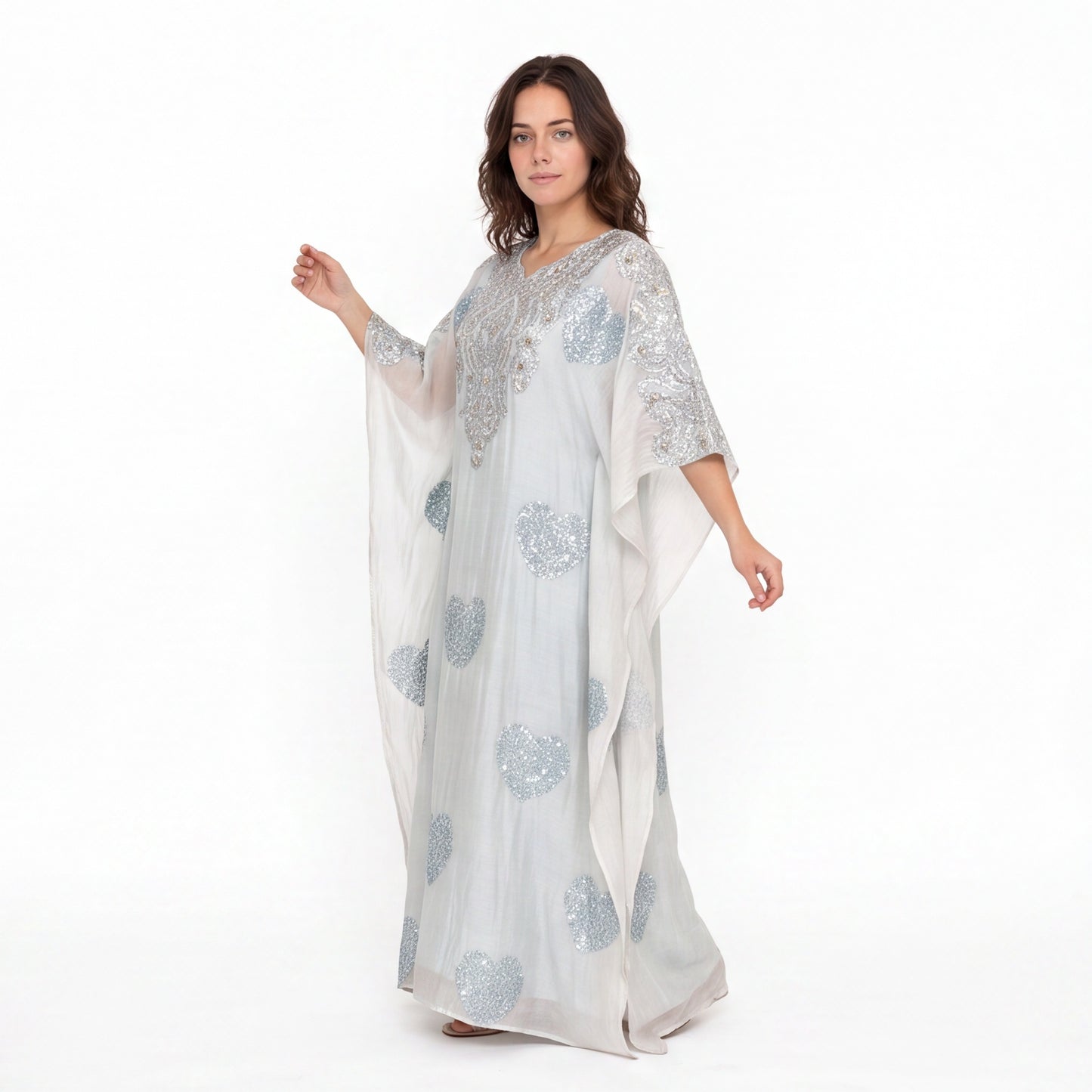 A woman modeling a long, flowing white caftan dress with silver sequin and bead embroidery, set against a plain white studio background.
