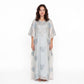 A woman modeling a long, flowing white caftan dress with silver-blue floral embroidery and beadwork, set against a plain white studio background.
