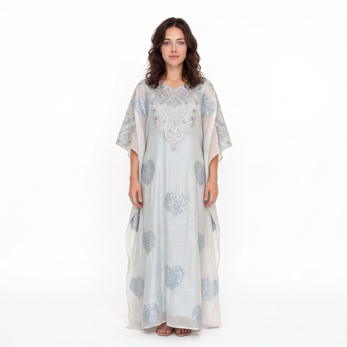 A woman modeling a long, flowing white caftan dress with silver-blue floral embroidery and beadwork, set against a plain white studio background.