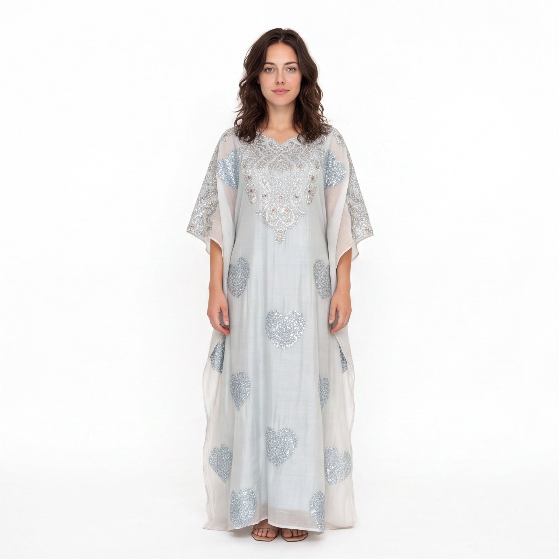A woman modeling a long, flowing white caftan dress with silver-blue floral embroidery and beadwork, set against a plain white studio background.