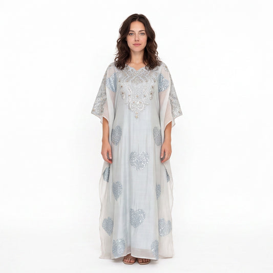 A woman modeling a long, flowing white caftan dress with silver-blue floral embroidery and beadwork, set against a plain white studio background.
