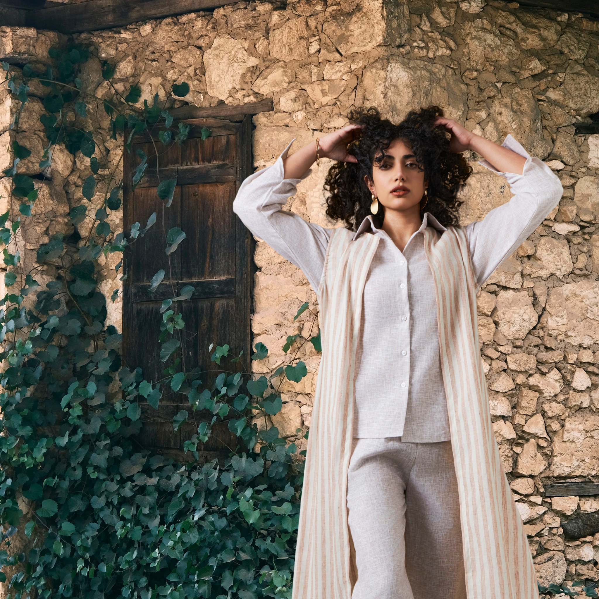 Woman in a white outfit standing against a stone wall with greenery