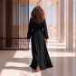 Back view of a woman walking in a floor-length black Marrakech kaftan with embroidered belt and sleeve cuffs, in a modern architectural setting.