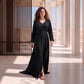 A woman with curly hair wearing a floor-length black Marrakech kaftan with tone-on-tone embroidery on the V-neckline, front panel and sleeves, tied with a matching belt, in a sunlit desert pavilion.