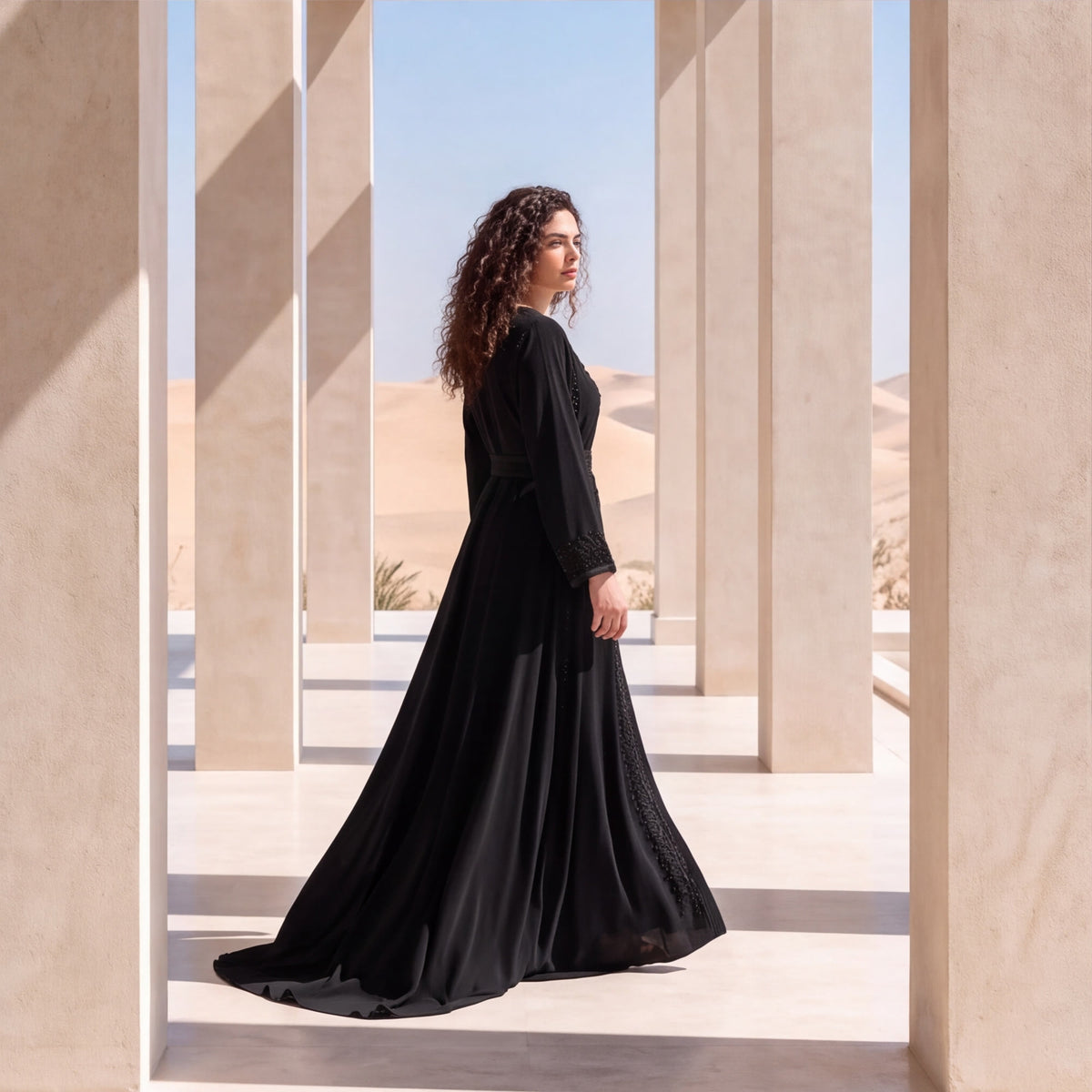 Side profile of a woman wearing a flowing black handmade Moroccan kaftan with a long train and embroidered sleeve cuff, standing between sandstone columns with desert dunes behind.