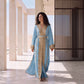 A woman wearing a floor-length sky blue Marrakech kaftan with gold and crystal embroidery on the neckline, front panel, sleeves and belt, walking through a sunlit desert pavilion.