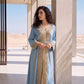 Half-body view of a woman in a sky blue Marrakech kaftan with gold and crystal embroidery on the V-neckline and a matching embroidered belt, standing beside a stone pillar.