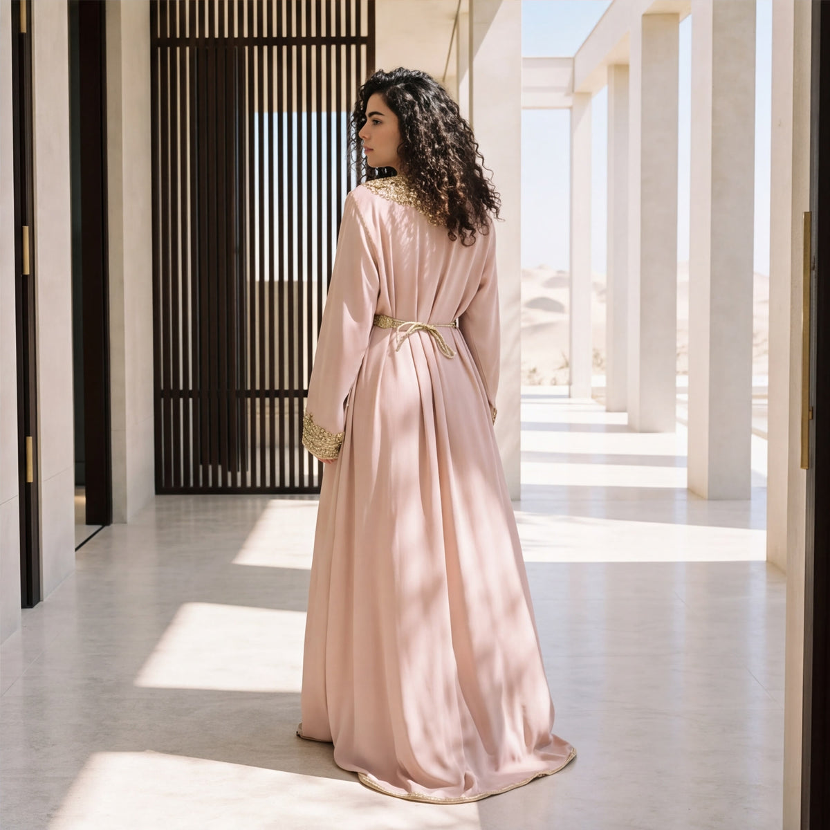 Back view of a woman wearing a floor-length blush pink handmade Moroccan kaftan with gold scroll trim.