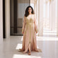 A woman wearing a floor-length blush pink kaftan with delicate gold scroll trim and a matching gold belt, standing in a sunlit architectural setting.