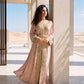 A woman wearing a dusty rose kaftan with gold beaded lace embroidery and a matching belt, in a Moroccan architectural setting.
