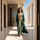 A woman wearing a deep emerald green floor-length kaftan with ornate gold filigree embroidery, standing in a sunlit architectural setting.