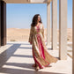 A woman wearing a long beige and pink floral caftan with a gold belt stands on a sunlit terrace with desert dunes and columns in the background.