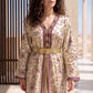 A woman wearing a beige embroidered kaftan robe with purple and gold floral patterns, a wide gold belt, and striped trim, photographed in a sunlit architectural setting.