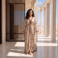 A woman stands in a sunlit architectural corridor wearing a long beige kaftan-style robe with an allover floral print, dark trim, and a gold belt.