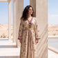 A woman wearing a long beige kaftan dress with purple floral patterns and gold trim, standing in a desert courtyard with columns.