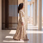 A woman stands in profile wearing a long beige kaftan dress with a small floral print and dark brown border details, set in a modern architectural corridor with columns and a desert landscape in the background.