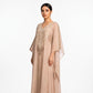 A woman modeling a long, flowing kaftan dress in a soft blush-beige color with intricate gold embroidery on the chest and sleeve edges, set against a plain white studio background.