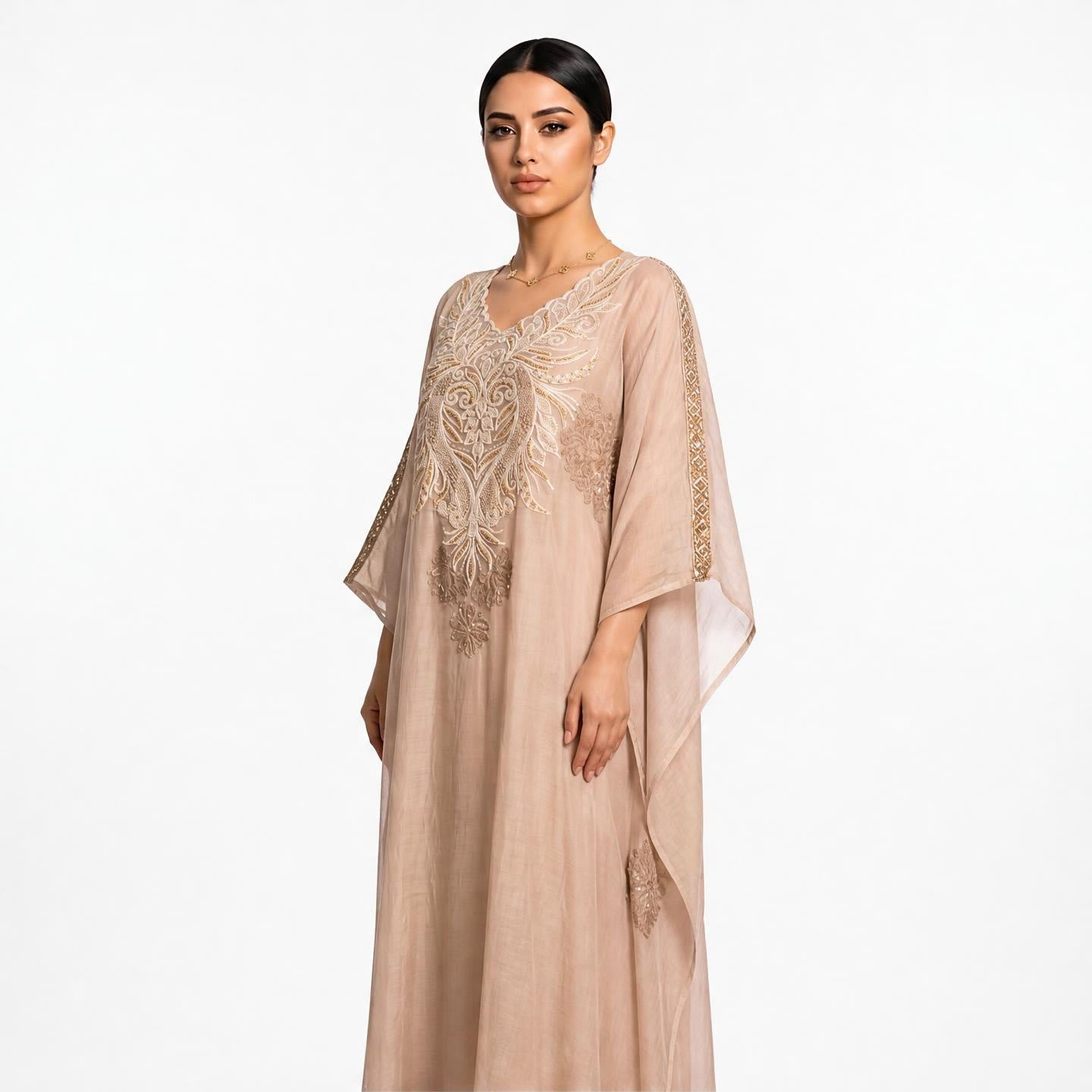 A woman modeling a long, flowing kaftan dress in a soft blush-beige color with intricate gold embroidery on the chest and sleeve edges, set against a plain white studio background.