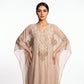 A woman modeling a beige kaftan dress with intricate gold embroidery and sheer sleeves, set against a plain white studio background.
