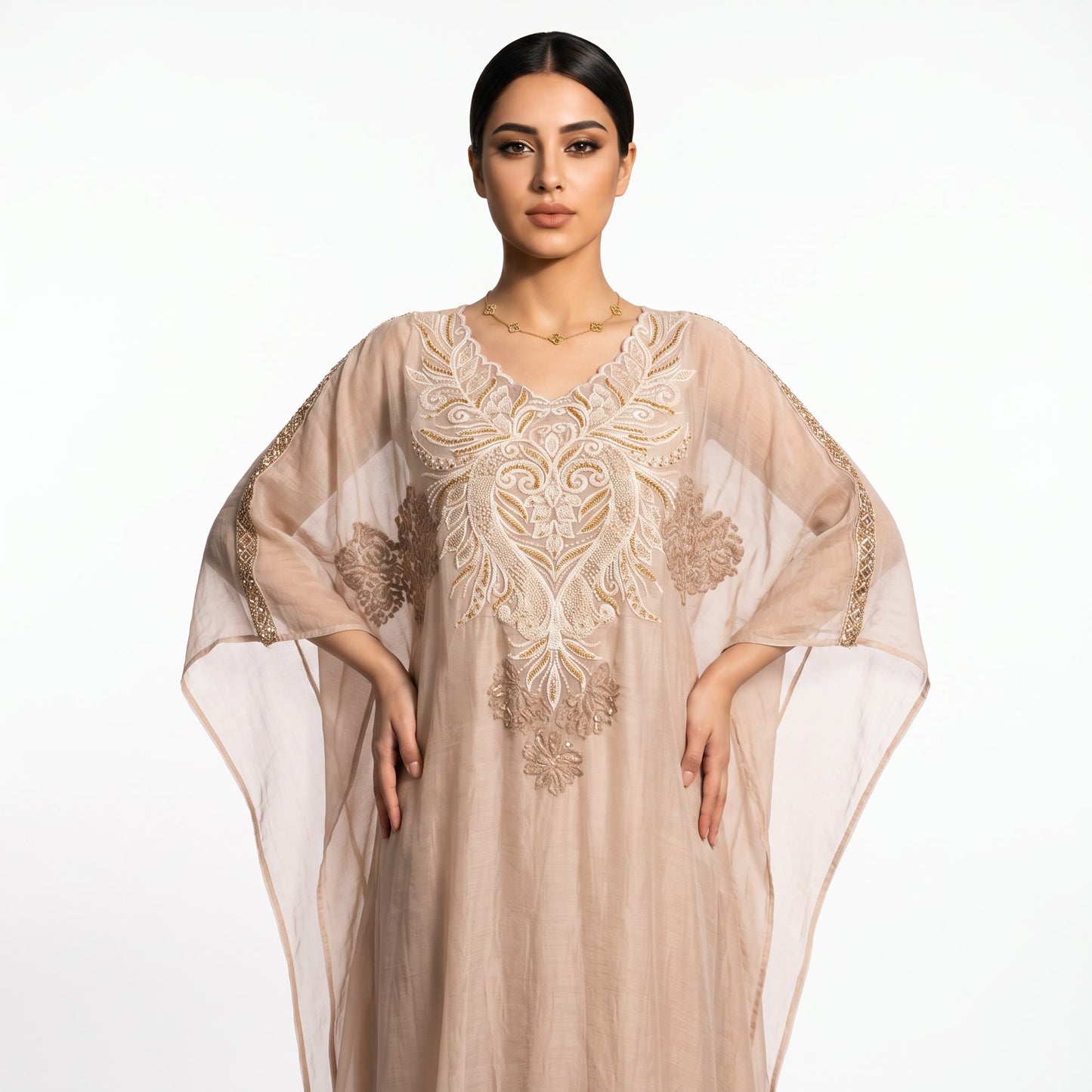 A woman modeling a beige kaftan dress with intricate gold embroidery and sheer sleeves, set against a plain white studio background.