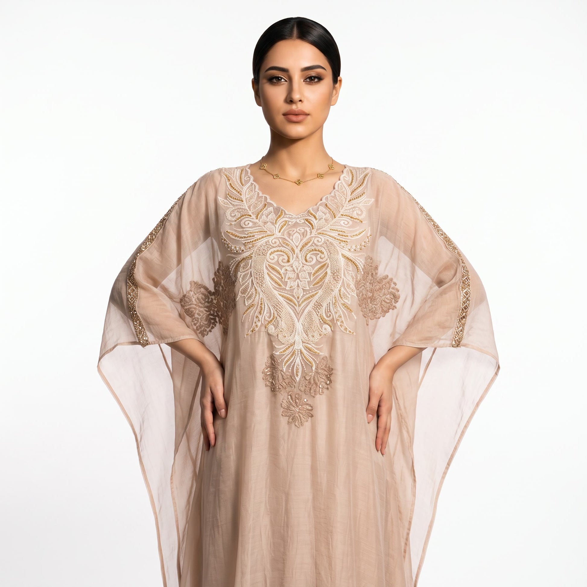 A woman modeling a beige kaftan dress with intricate gold embroidery and sheer sleeves, set against a plain white studio background.