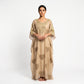 A woman stands barefoot against a plain white studio background wearing a loose, floor-length beige kaftan dress with gold embroidery and heart motifs.