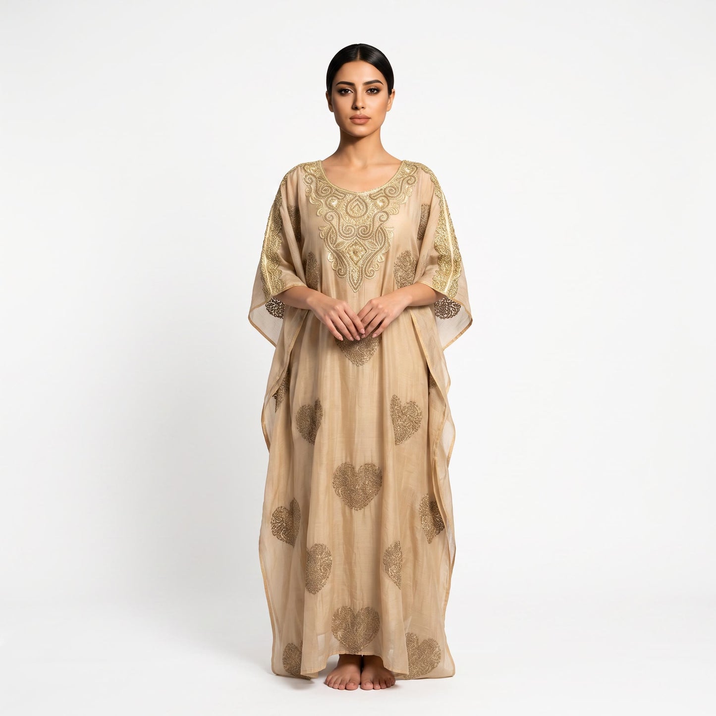A woman stands barefoot against a plain white studio background wearing a loose, floor-length beige kaftan dress with gold embroidery and heart motifs.