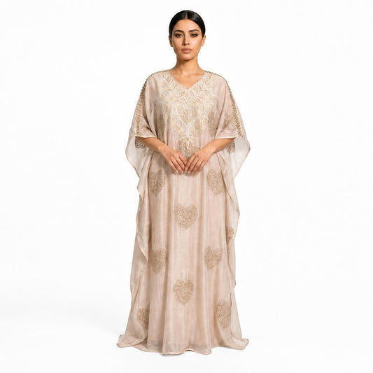 A woman modeling a floor-length beige caftan dress with gold embroidery and sheer draped sleeves against a plain white background.