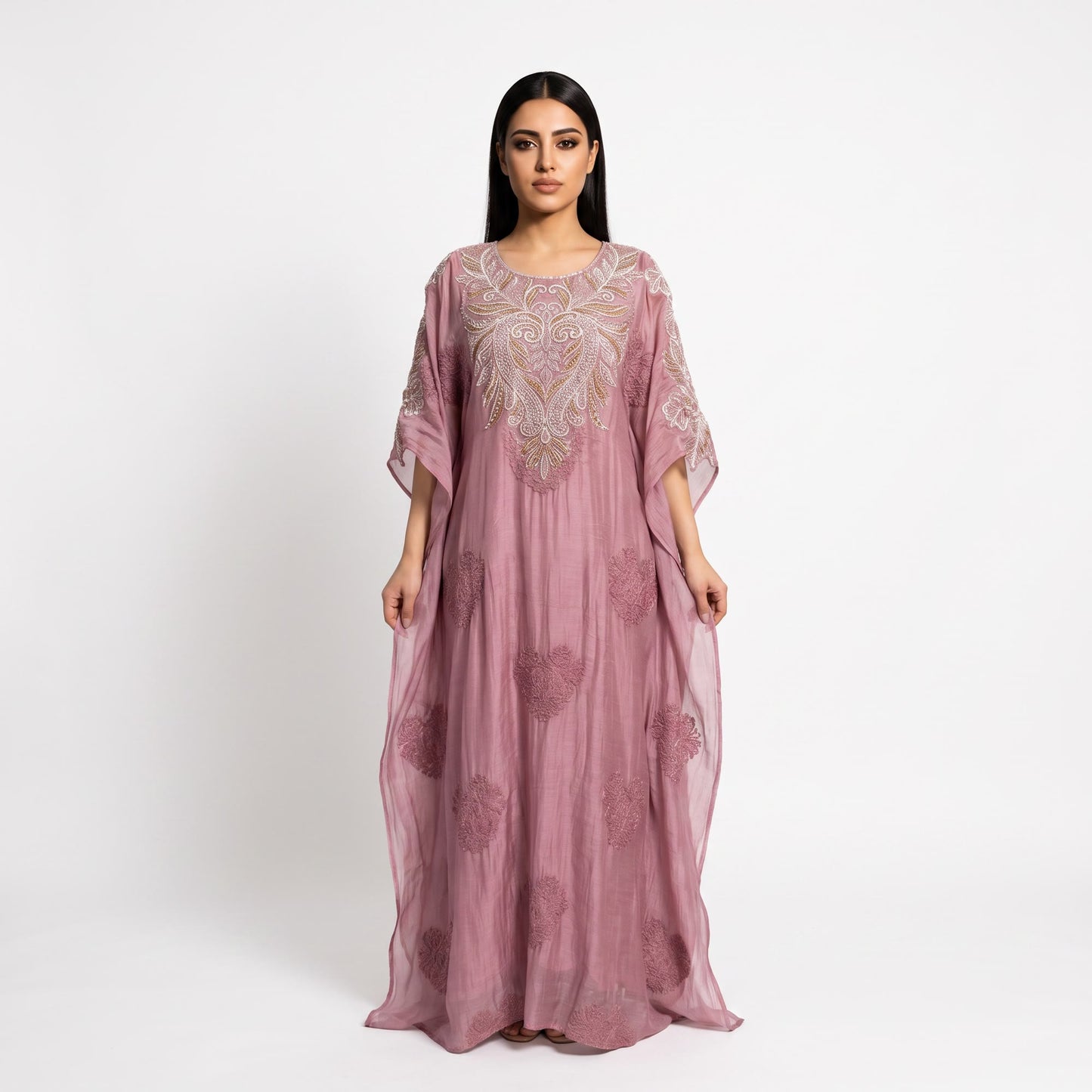 A woman modeling a dusty pink kaftan dress with intricate silver and gold embroidery on the bodice and scattered floral patterns on the flowing skirt, set against a plain white studio background.