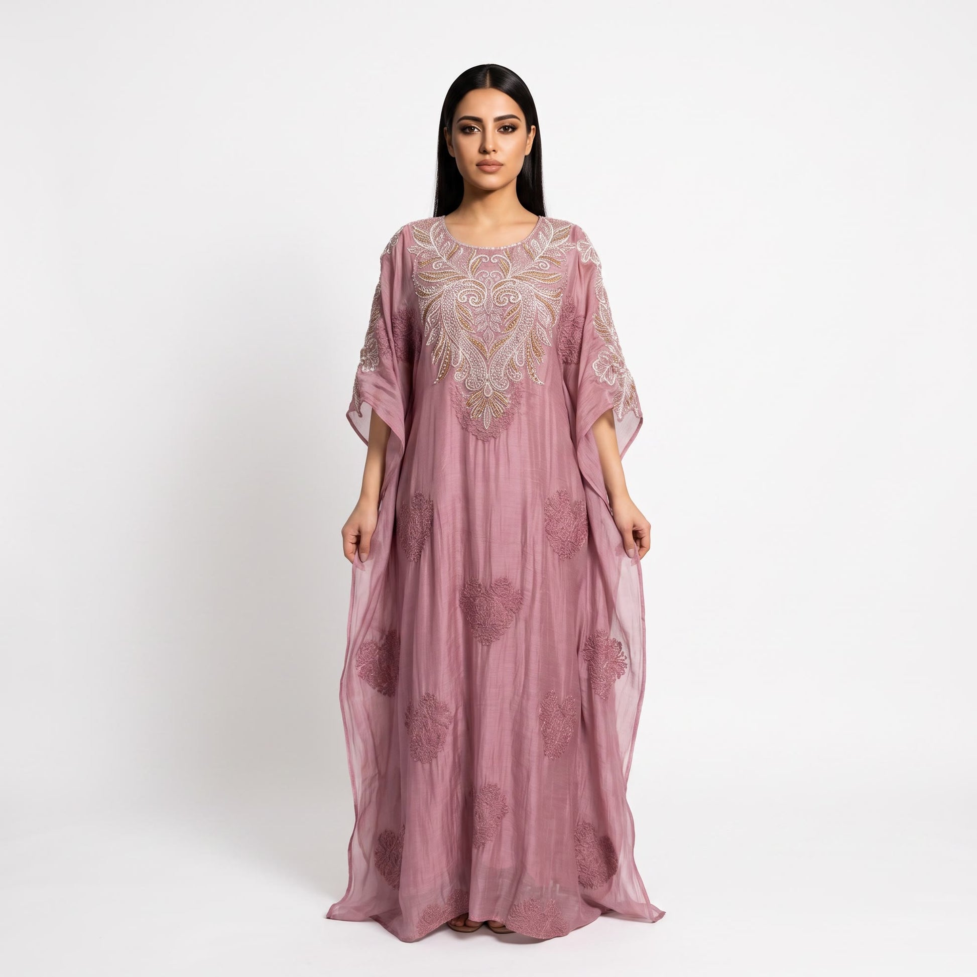 A woman modeling a dusty pink kaftan dress with intricate silver and gold embroidery on the bodice and scattered floral patterns on the flowing skirt, set against a plain white studio background.