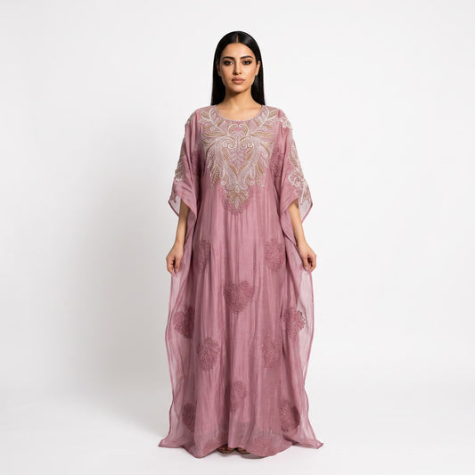 A woman modeling a dusty pink kaftan dress with intricate silver and gold embroidery on the bodice and scattered floral patterns on the flowing skirt, set against a plain white studio background.