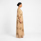 A woman in profile wearing a floor-length beige kaftan dress with gold floral patterns, photographed against a clean white studio background.