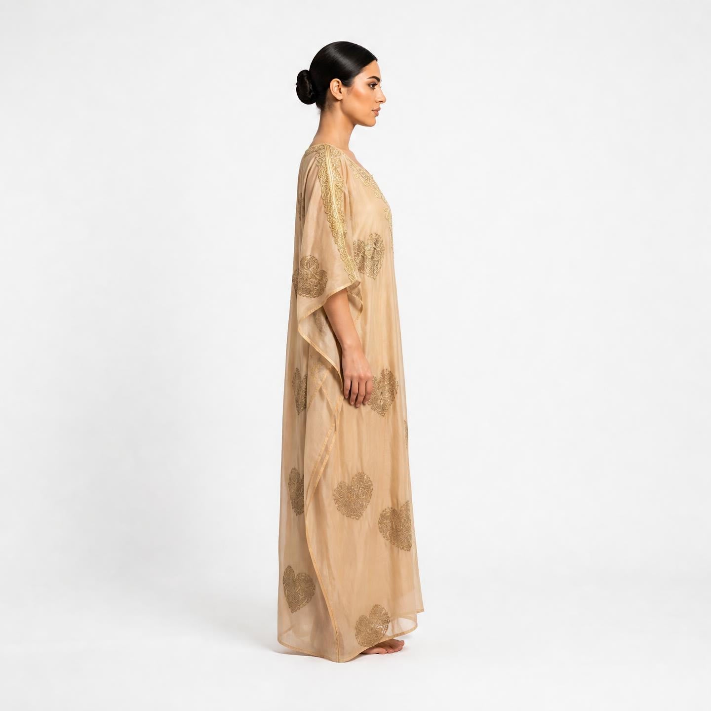 A woman in profile wearing a floor-length beige kaftan dress with gold floral patterns, photographed against a clean white studio background.