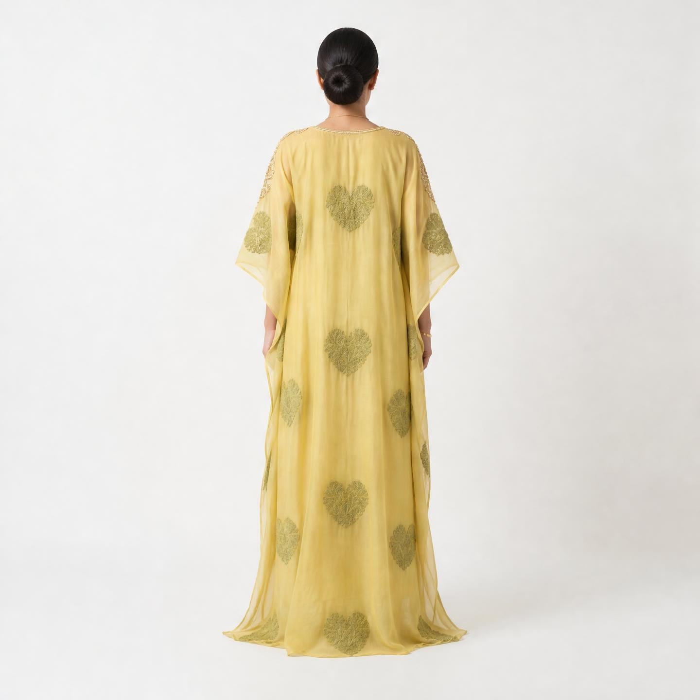 A woman viewed from the back wearing a flowing yellow kaftan dress with olive green heart motifs, set against a plain light gray studio background.