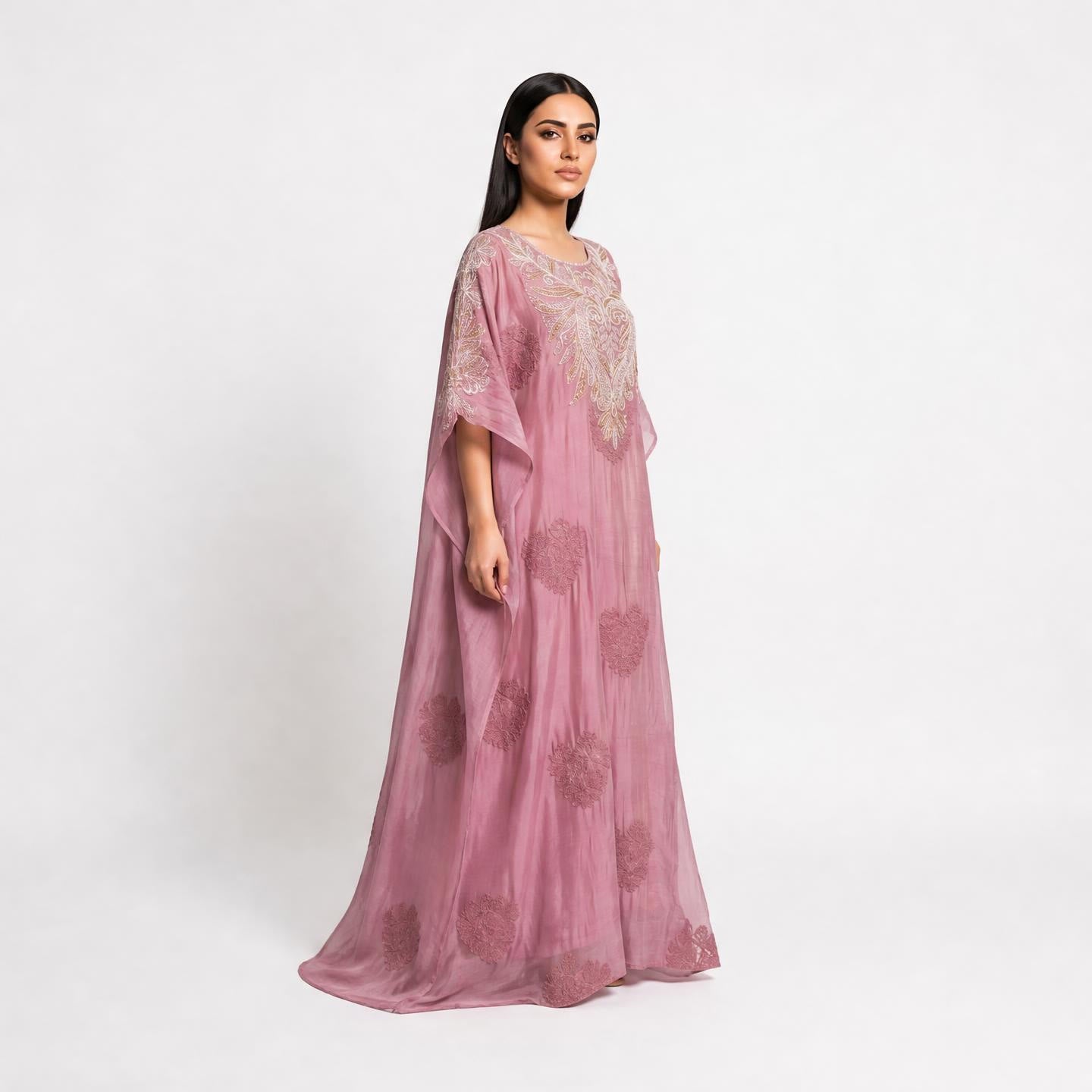 A woman modeling a long, flowing dusty pink kaftan dress with intricate floral embroidery and sheer sleeves, photographed against a plain white studio background.